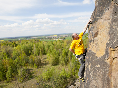 Ein Mann klettert einen Steinbruch hinauf, im Hintergrund Natur und eine Ortschaft, Aktiv, Klettern, Natur