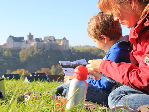 Eine Frau mit Kind sitzt mit einer Broschüre in der Hand auf einer Wiese, im Hintergrund erhebt sich Burg Mildenstein, Auflug, Familie,