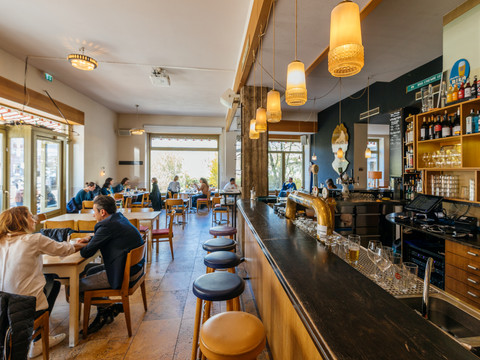 Blick auf die Bar und den vorderen Gastraum des Café Cantona mit gemütlichen Holzmöbeln und einer großen Fensterfront