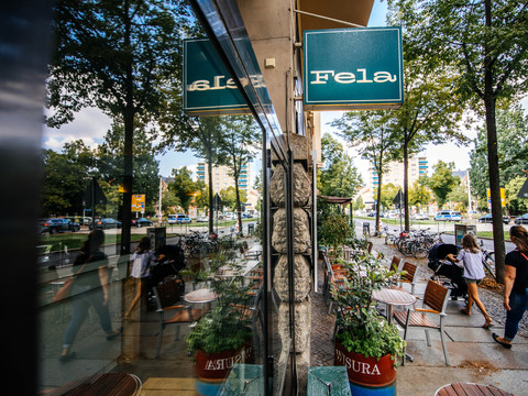 Blick entlang der Fassade und des großen Schaufensters des Fela Restaurants, auf das blaue Geschäftsschild, den Straßenzug und die Spiegelung im Fester; an einem Sommertag, freisitz, restaurant, grastronomie