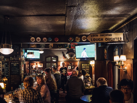 Blick in das gut gefüllte Irish Pub; Gäste sitzen an der Bar, schauen eine Fußballübertragung oder unterhalten sich im dunklen Lokal, kneipe, pub, gastronomie
