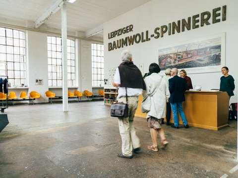 Blick in den Eingangsbereich/ das Foyer der Spinnerei mit Besuchern und Mitarbeitern; an der weißen Wand befindet sich der Schriftzug der Baumwollspinnerei, davor steht ein gelber Tresen und Sitzgelegenheiten, Leipzig und Region