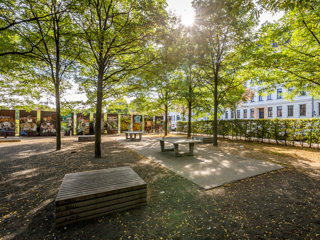 Blick auf die von Bäumen umgebenen und schattigen Tischtennisplatten im Lene-Voigt-Park; im Hintergrund: Häuserfassaden und Begrenzungen mit Graffiti