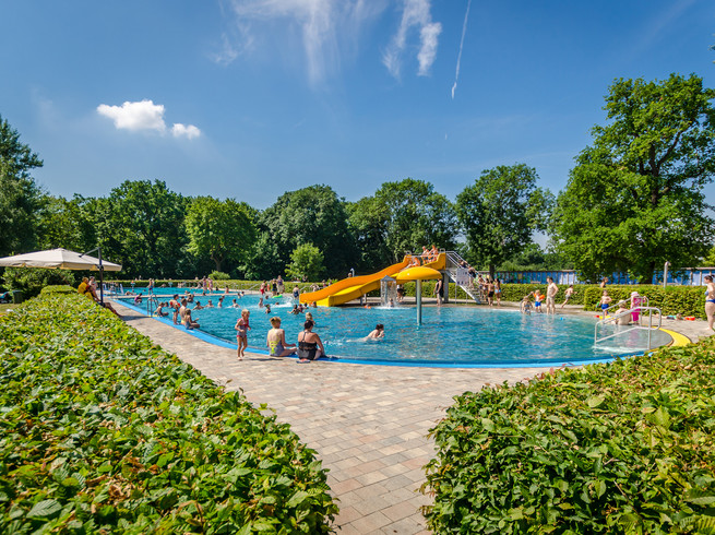 Blick auf das Hauptschwimmbecken im Schreberbad, mit gelber Rutsche und vielen Badegästen an einem Sommertag