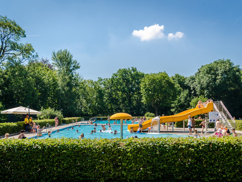 Blick auf das Hauptschwimmbecken im Schreberbad, mit gelber Rutsche und vielen Badegästen an einem Sommertag